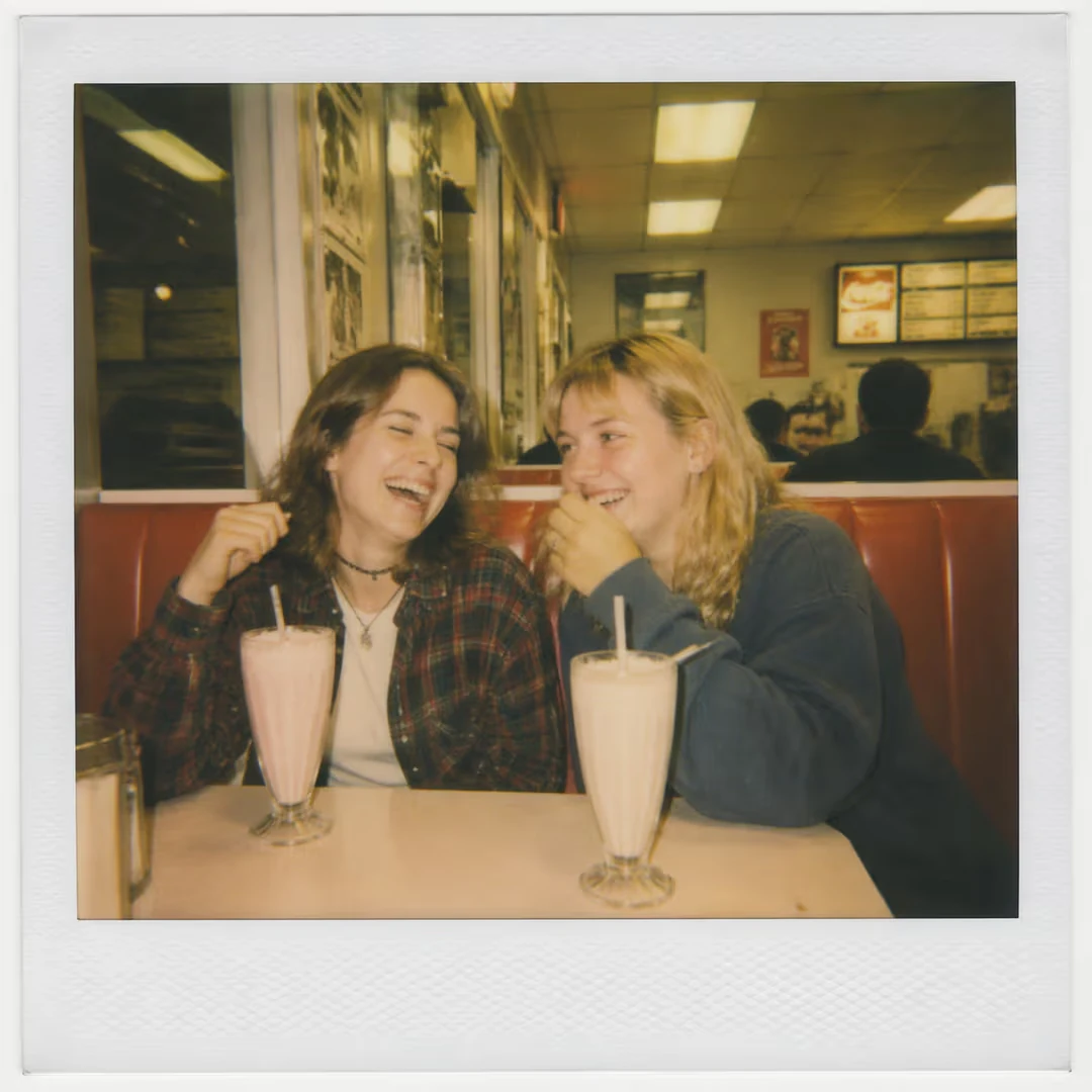 A candid Polaroid-style photograph of two friends laughing at a 1990s diner booth. Slightly washed-out colors with a war